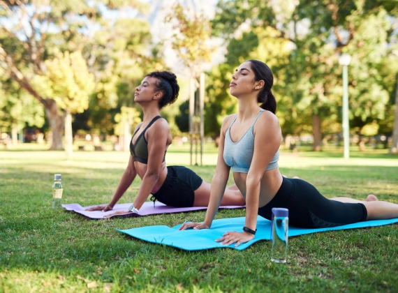 Two women do yoga on mats in a grassy, sunny park.
