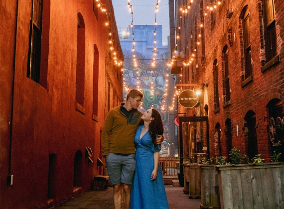 A smiling couple in elegant attire, looking at each other under illuminated strings lights in an alley