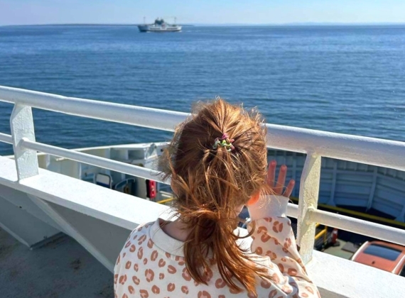 A young girl observes a boat in the distance from the deck of a ferry.