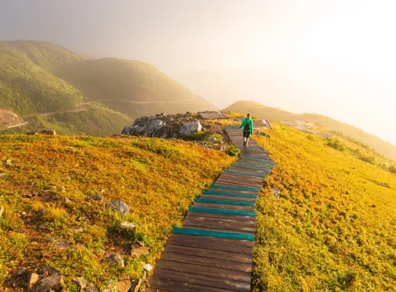 A person walking along a boardwalk trail on top of a cliff overlooking mountains and coast line at sunset.