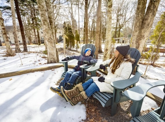 A woman and child sitting in Muskoka chairs, laughing in a snowy wooded area.
