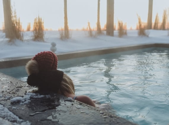 Two people wearing winter hats watching a sunset in a hot tub on a snowy evening 