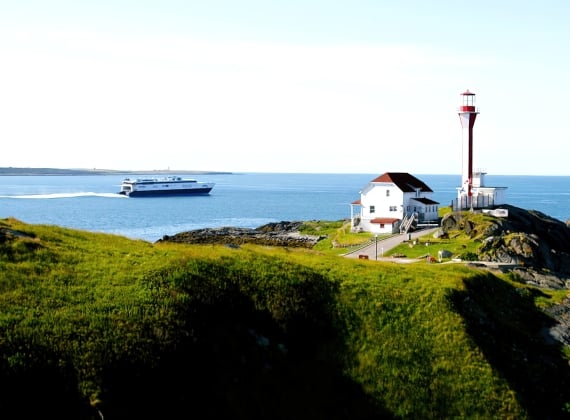 A small building and lighthouse on green, rocky cliff side overlooking the ocean with a large ferry in the background on a sunny day.