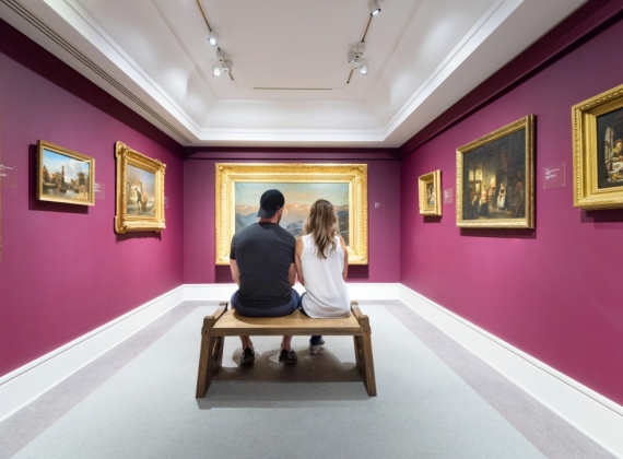 A couple sits on an art gallery bench and observes a painting