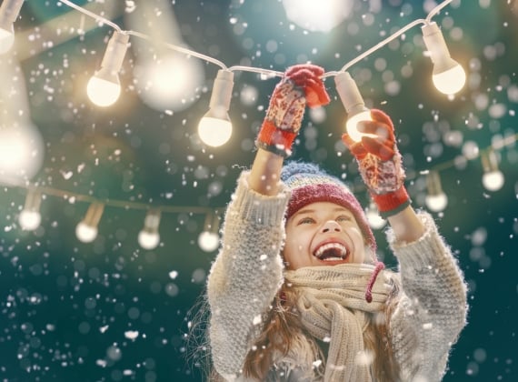 Child smiling and playing with string globe lights in a light snowfall.