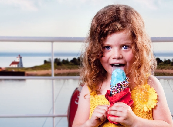 Young child wearing a yellow dress eating a blue ice cream cone with sunny seaside background