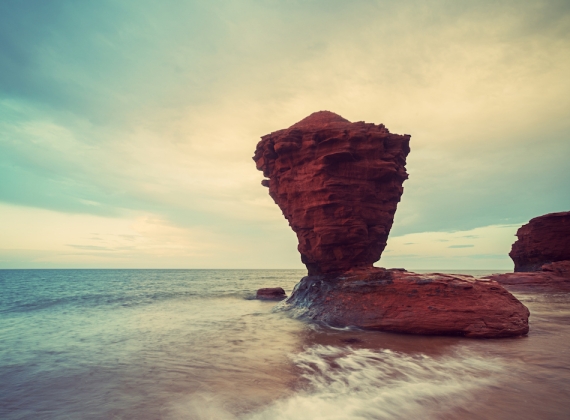 A large red sandstone rock near the ocean, looking as if it is balancing on the rock it stands on.