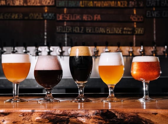 5 different beers in a range of styles of glasses on a wooden bar top.