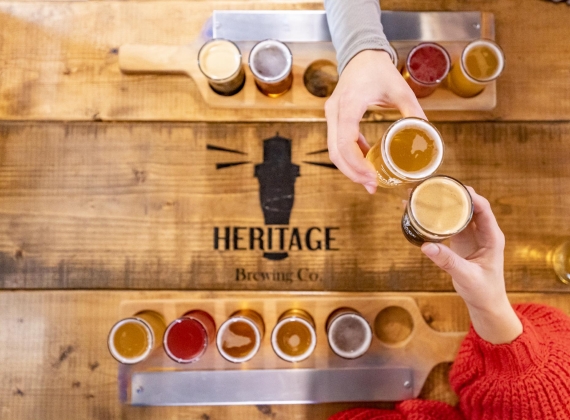 An overhead view of two people clinking small glasses of beer, each with a flight of beer tasters on a wooden table branded with Heritage Brewing Co. label 