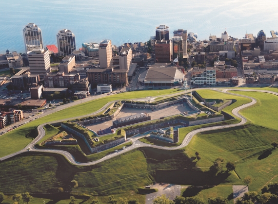An arial view of Halifax with Citadel Hill (a citadel in the shape of a star on top of a green hill) in the foreground, buildings and the harbour in the background.