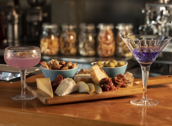 A pink cocktail, a purple cocktail with a charcuterie board of various cheeses, cured meats, nuts and olives on a wooden bar top.