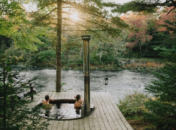 Two people in a hot tub in the forest, overlooking a river at sunset.