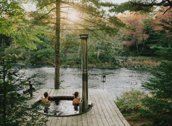 Two people in a hot tub in the forest, overlooking a river at sunset.