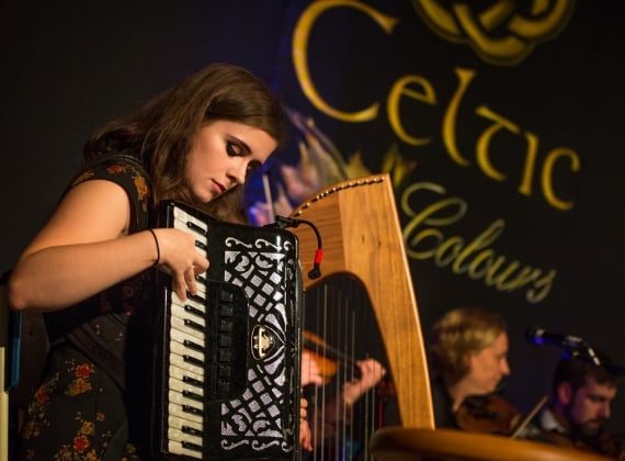 A woman playing accordion on stage with a harpist and fiddlers in the background.