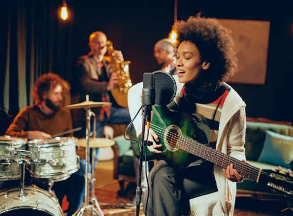 A woman playing acoustic guitar and singing with a drummer and a saxophonist in the background.