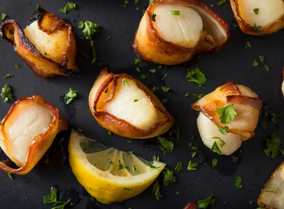 Overhead shot of bacon wrapped scallops with lemon and parsley garnish on a black plate.
