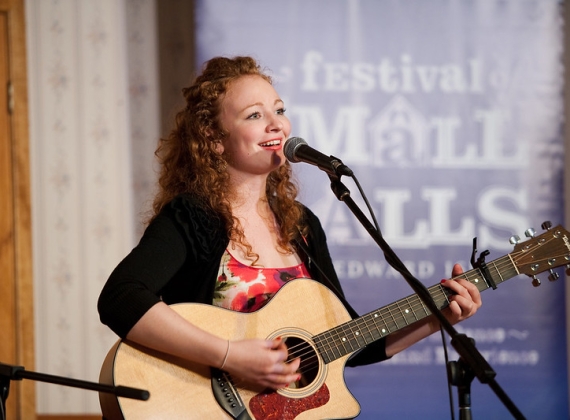 A woman sings into a microphone and plays acoustic guitar on stage