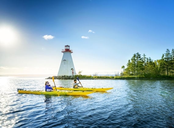 Two people in sea kayaks on calm water with a lighthouse and trees in the background.