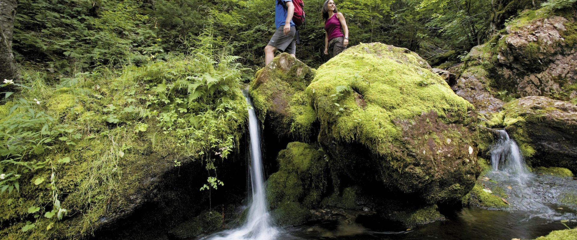 Two hikers pass by a small waterfall in a lush environment