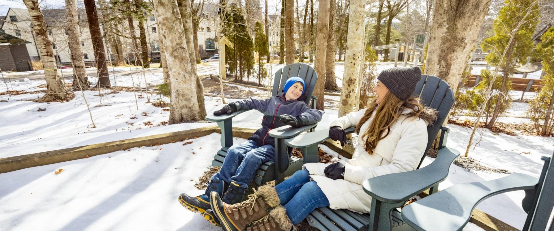 A woman and child sitting in Muskoka chairs, laughing in a snowy wooded area.