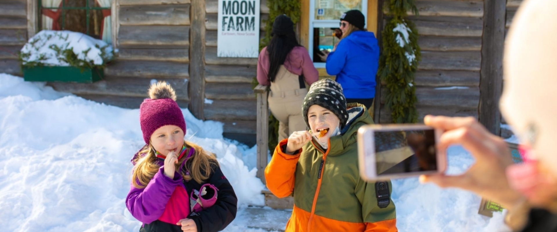 Two children have their picture taken while they eat maple syrup off of popsicle sticks in front of a wooden cabin in the winter.