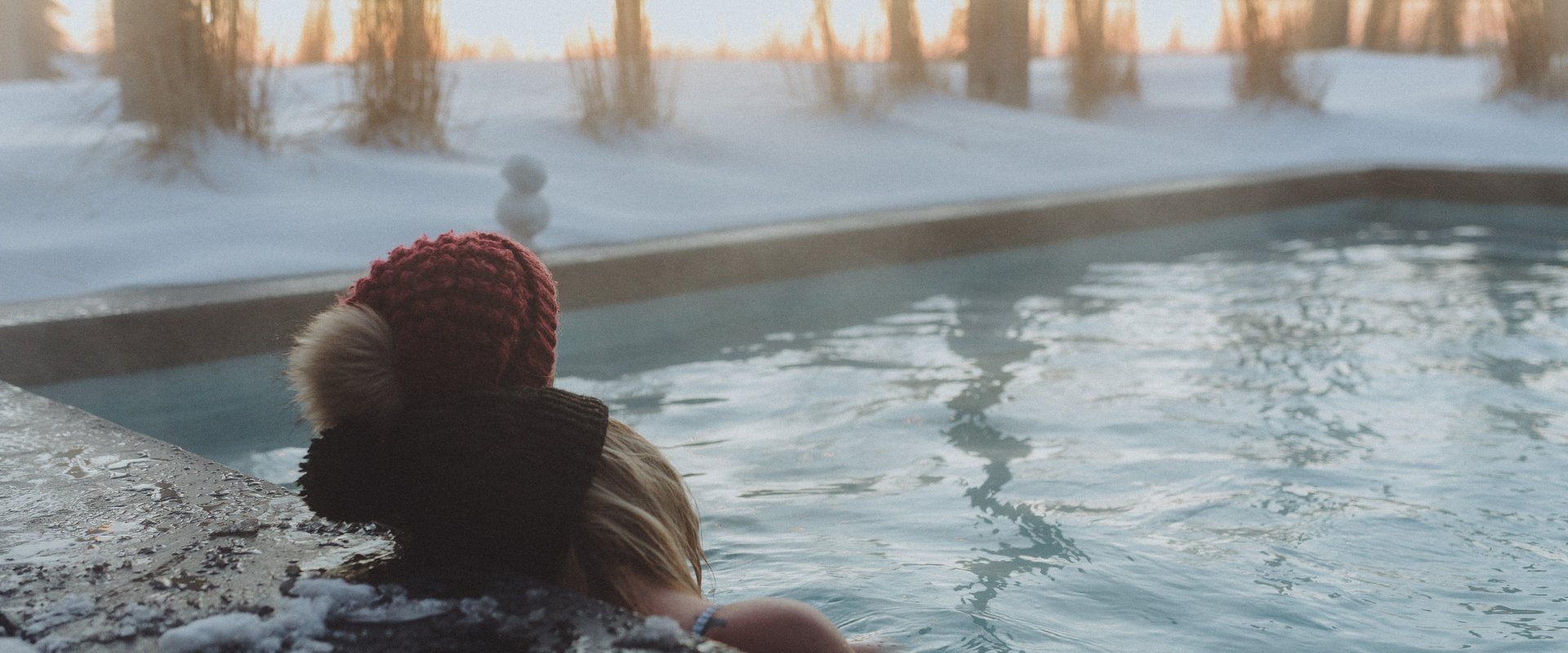 Two people wearing winter hats watching a sunset in a hot tub on a snowy evening 