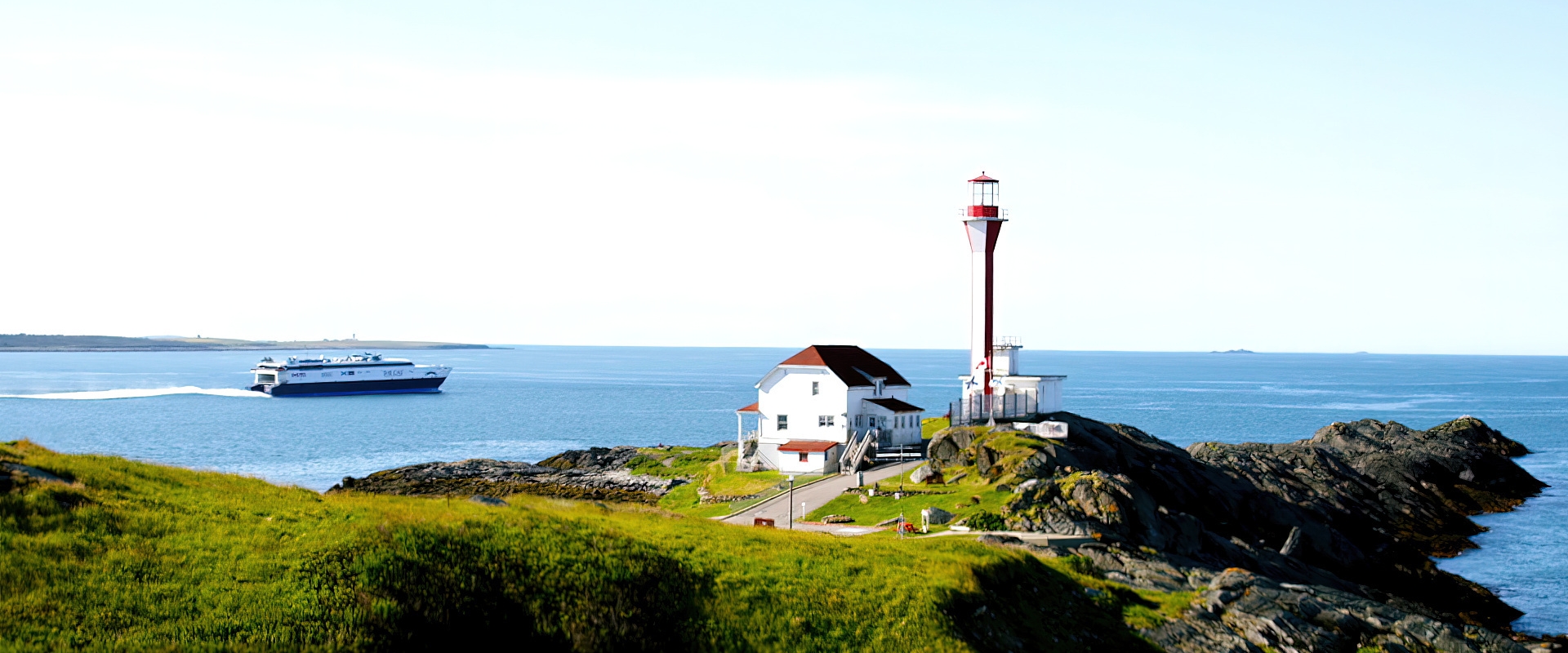 A small building and lighthouse on green, rocky cliff side overlooking the ocean with a large ferry in the background on a sunny day.