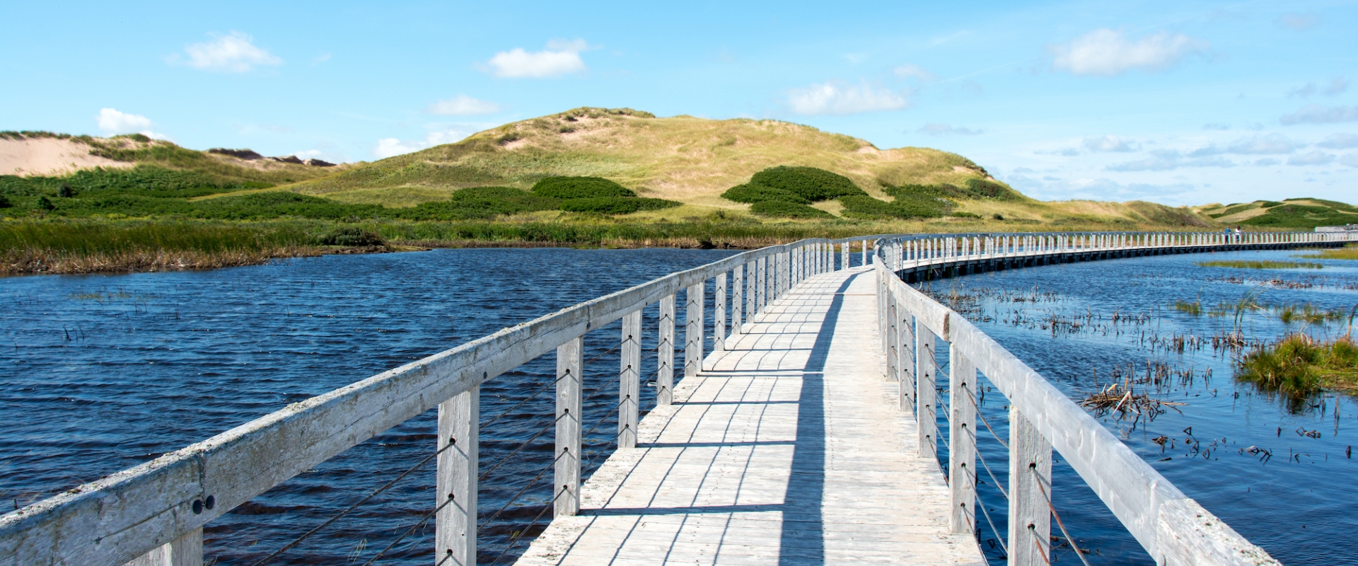 A wooden footbridge across calm blue water with rolling green hills in the background on a sunny day.