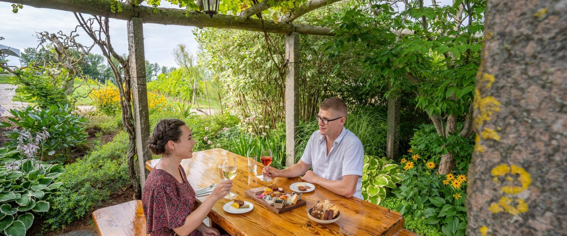 Two people enjoying lunch and wine at a picnic table surrounded by plants and greenery.